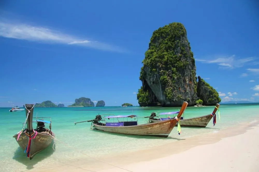 Longtail boats on a white beach below a limestone rock in Thailand